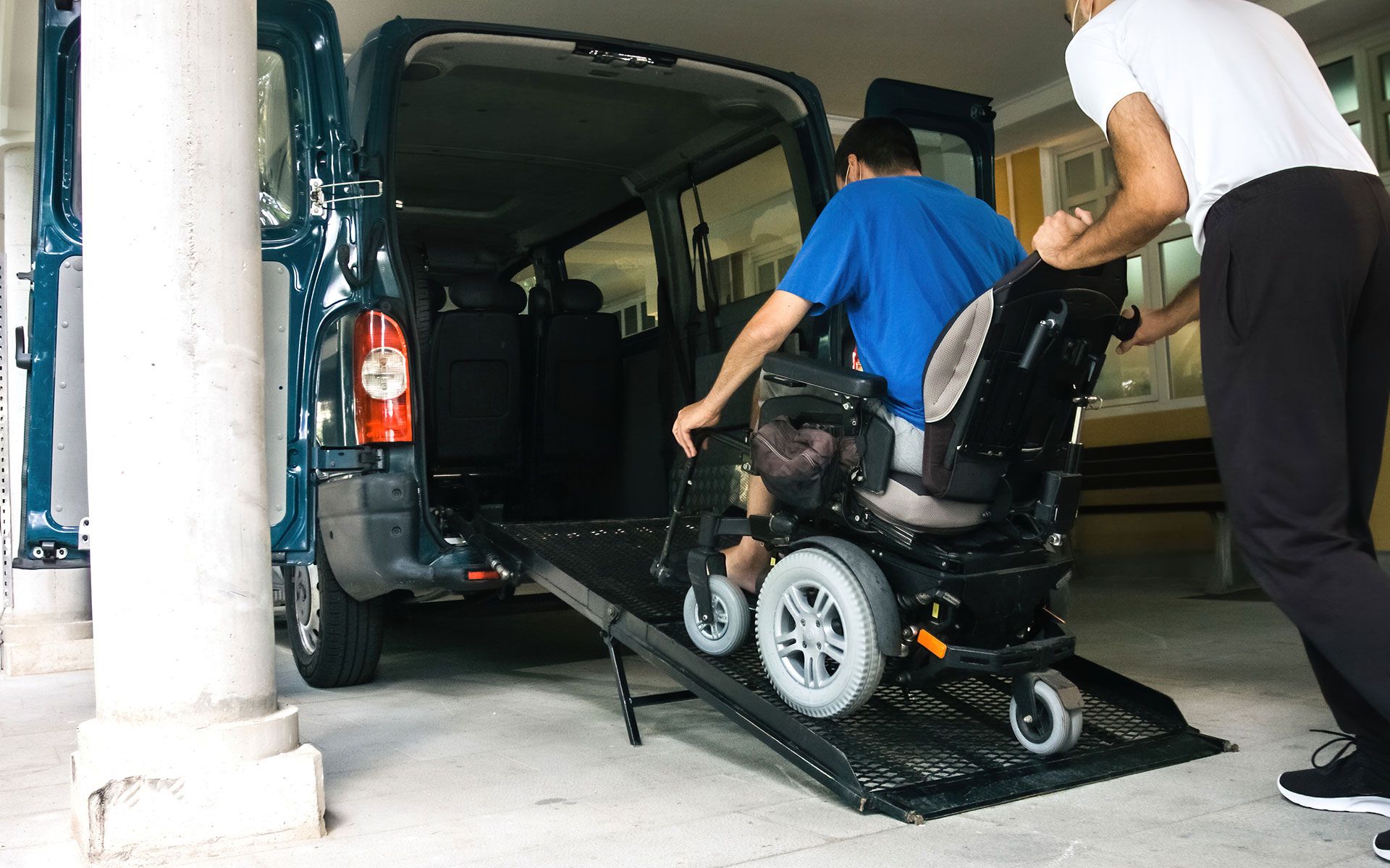 Man in wheelchair being pushed up a ramp into a dark blue van by another man.