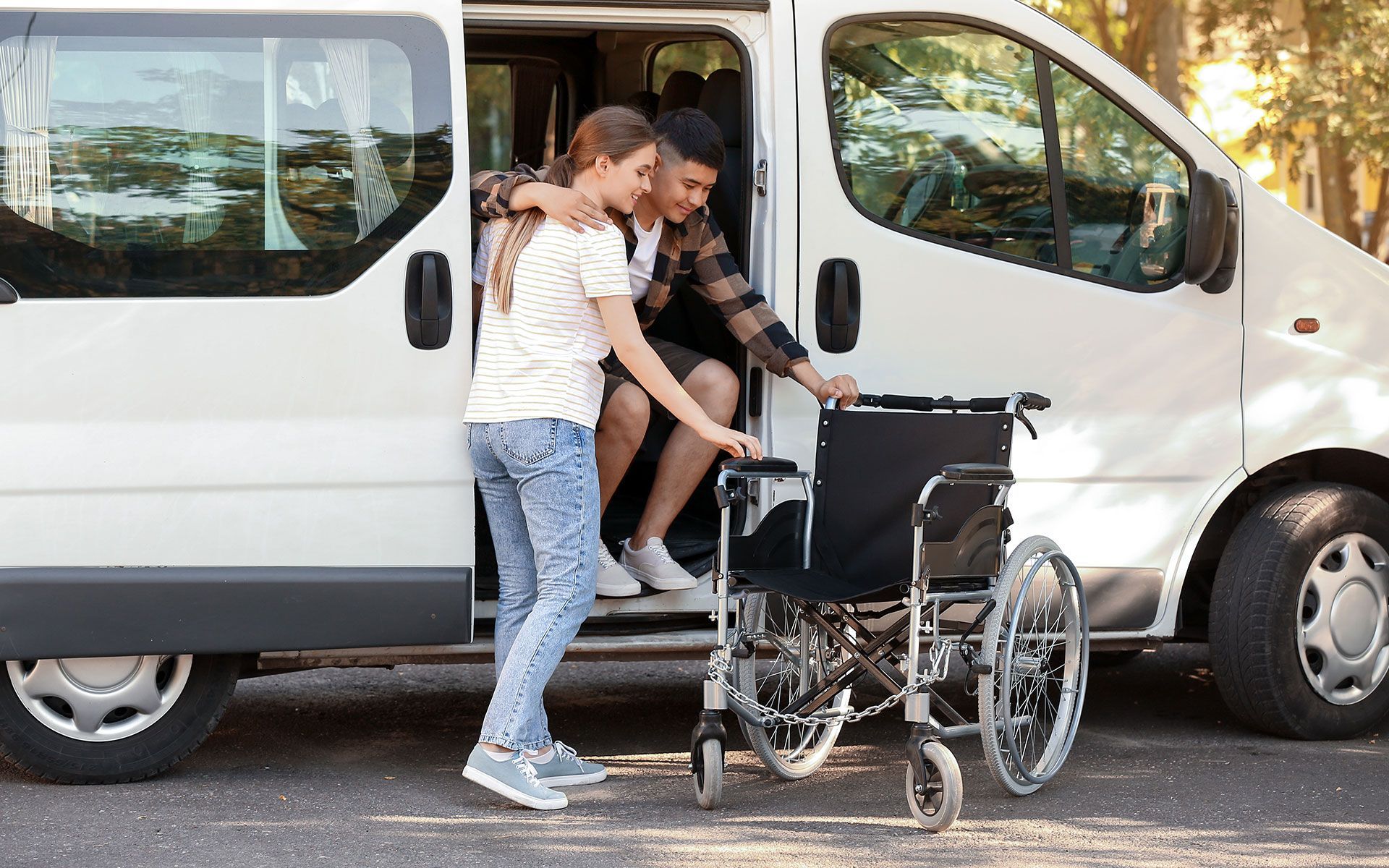 Two people helping a person with a disability exit a white van with a wheelchair on the ground.