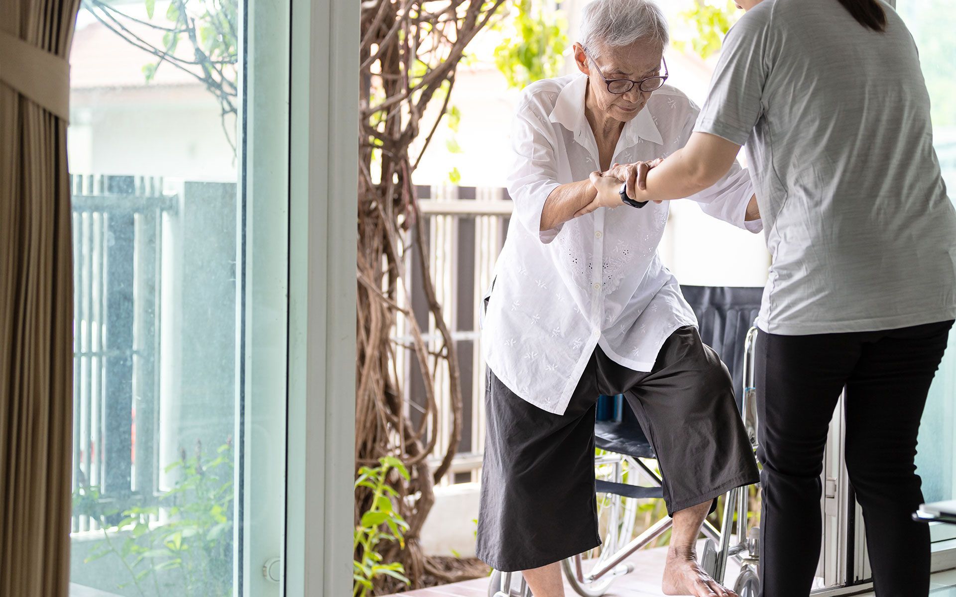 Elderly woman with help from caregiver, standing up from a wheelchair near a window.