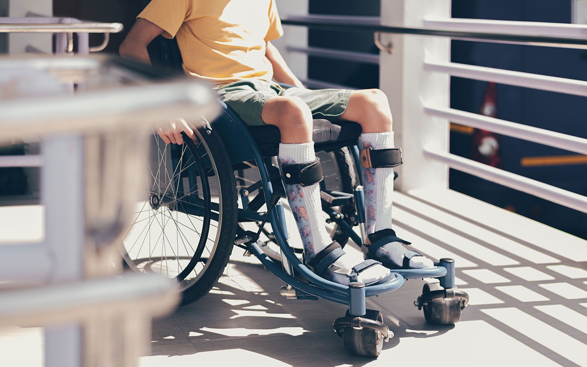 Person in a wheelchair on a ramp; wearing leg braces and a yellow shirt; outdoor setting.