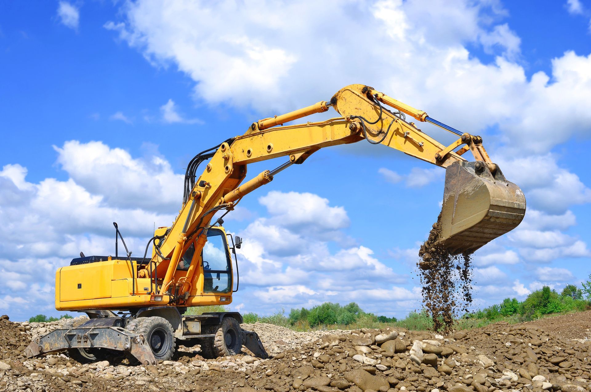 Un escavatore giallo scarica terra in un cantiere sotto un cielo azzurro.
