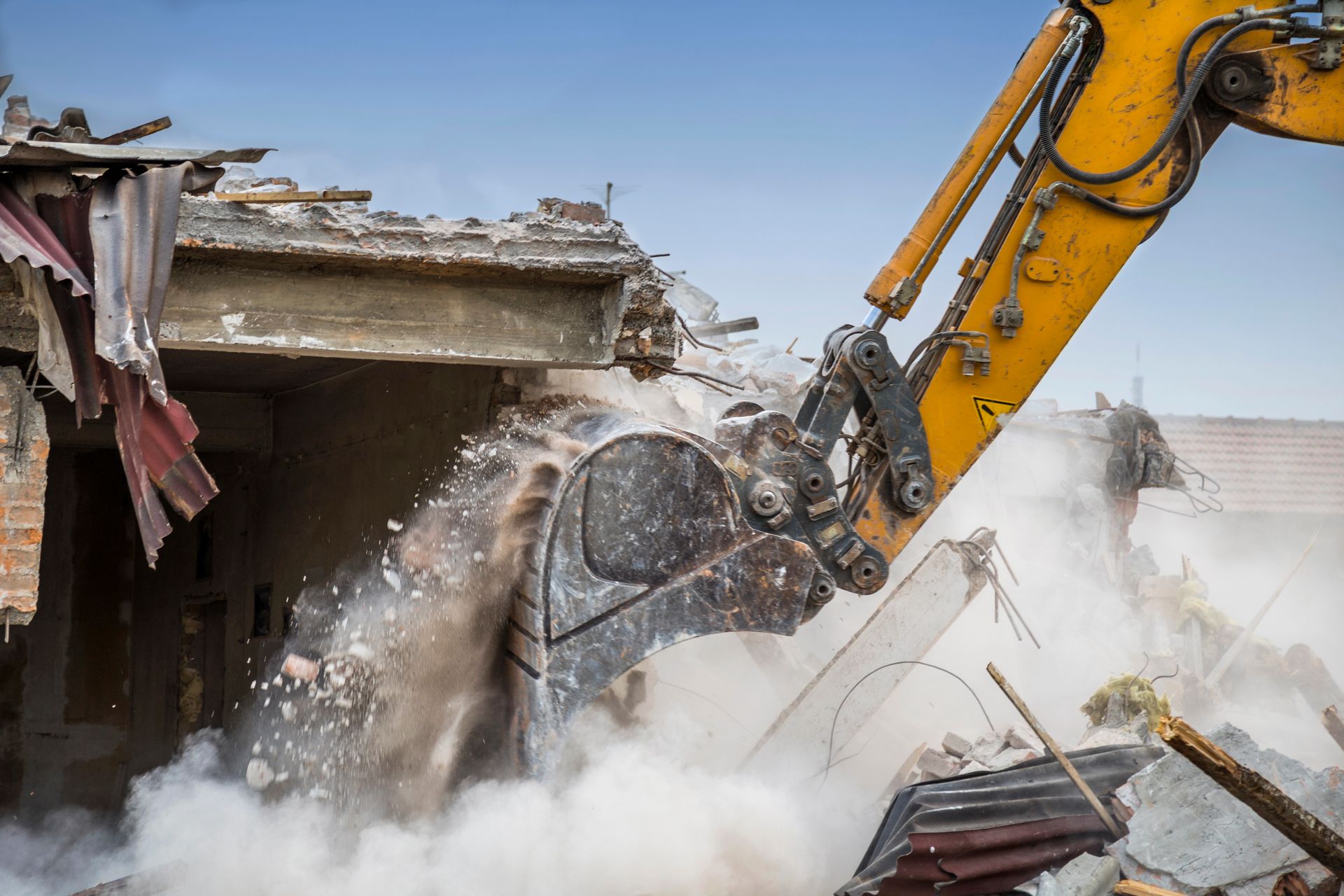 Un escavatore giallo sta demolendo un edificio in cemento, sollevando polvere e detriti.