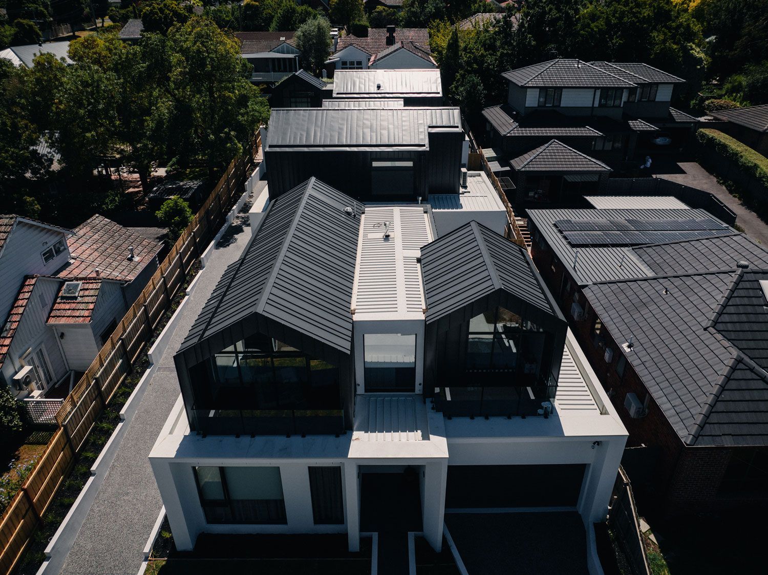 Aerial view of a modern white house with a dark gray roof, surrounded by trees and other houses.