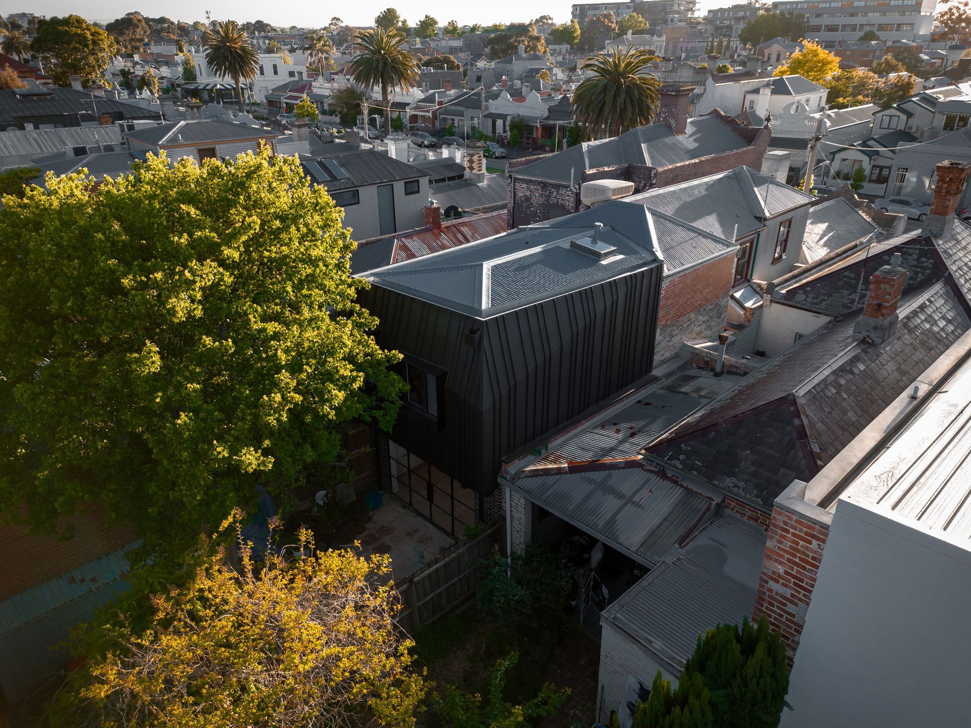 Aerial view of a residential neighborhood with dark wooden modern home contrasting older buildings, trees, and rooftops.
