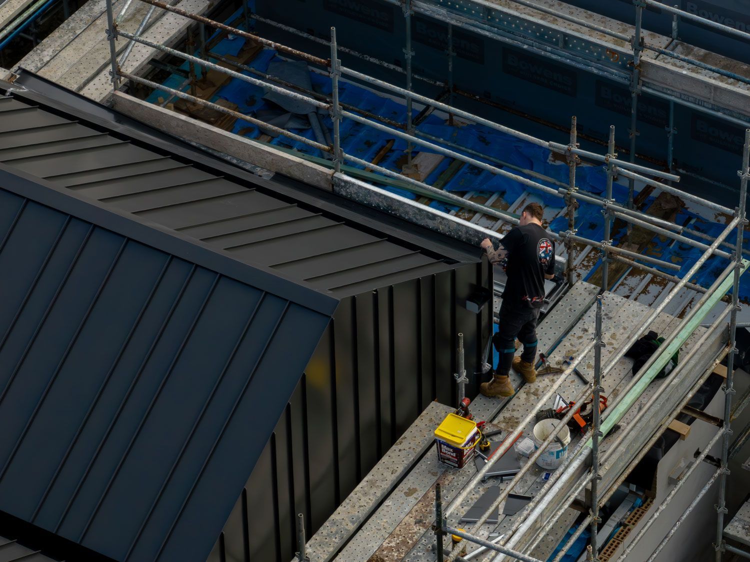 A construction worker on scaffolding near a building roof. He's wearing a black uniform, working with tools.