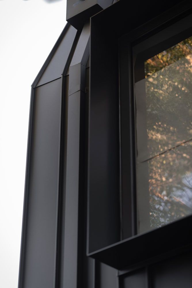 Close-up of a modern building's corner featuring black metal cladding, a window, and the reflection of trees.
