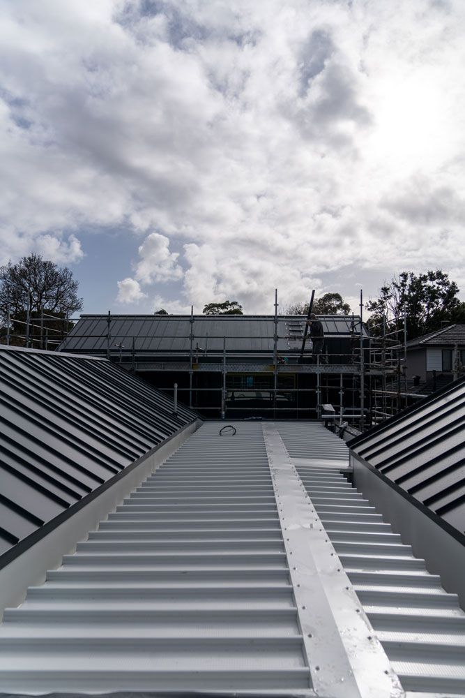 A newly constructed corrugated metal roof, with scaffolding and partially built house in the background under a cloudy sky.
