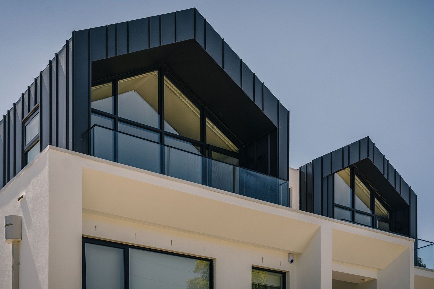 Modern white building with black angular roof, glass balconies, and windows under a blue sky.