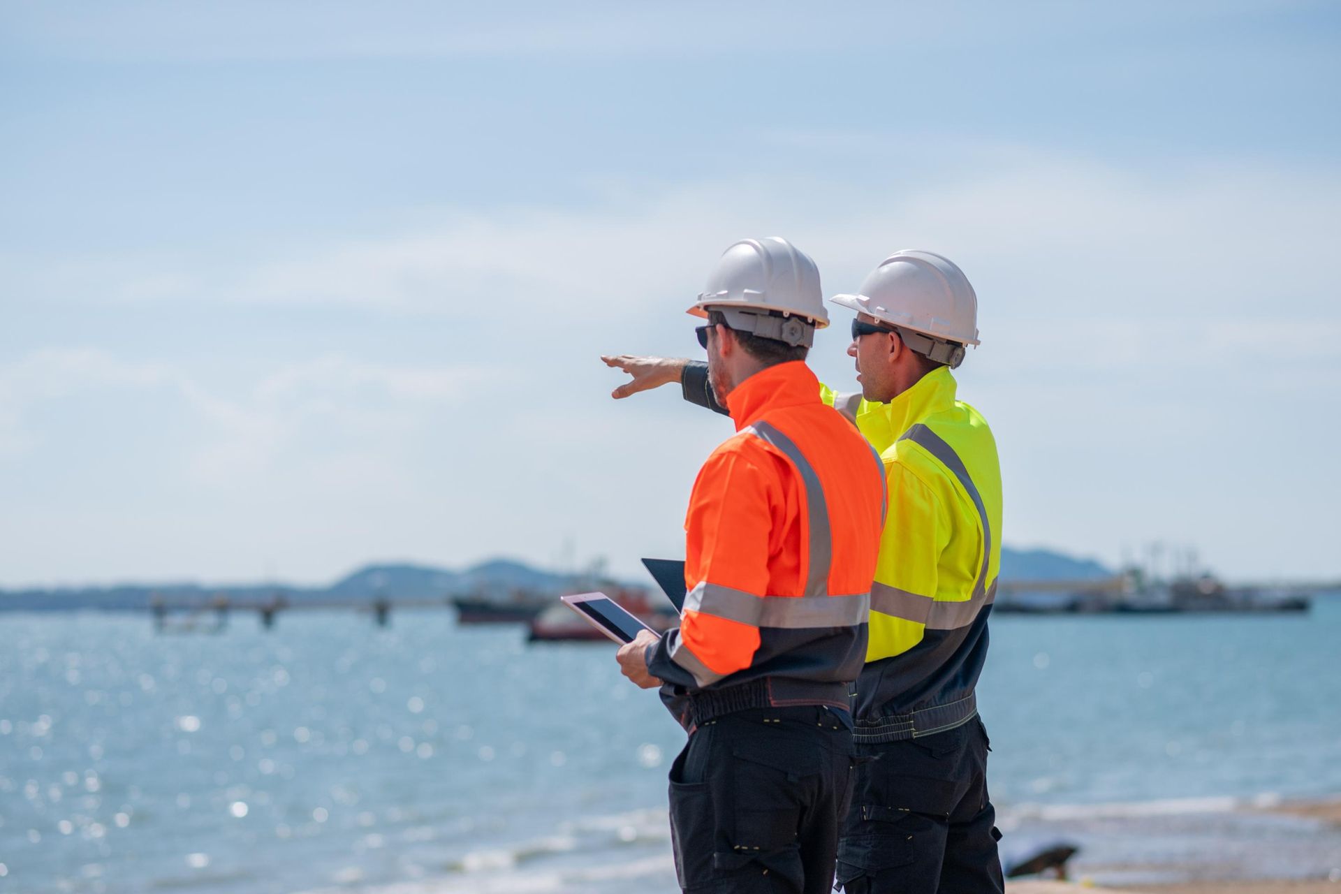 Two construction workers are standing on a beach looking at the ocean.