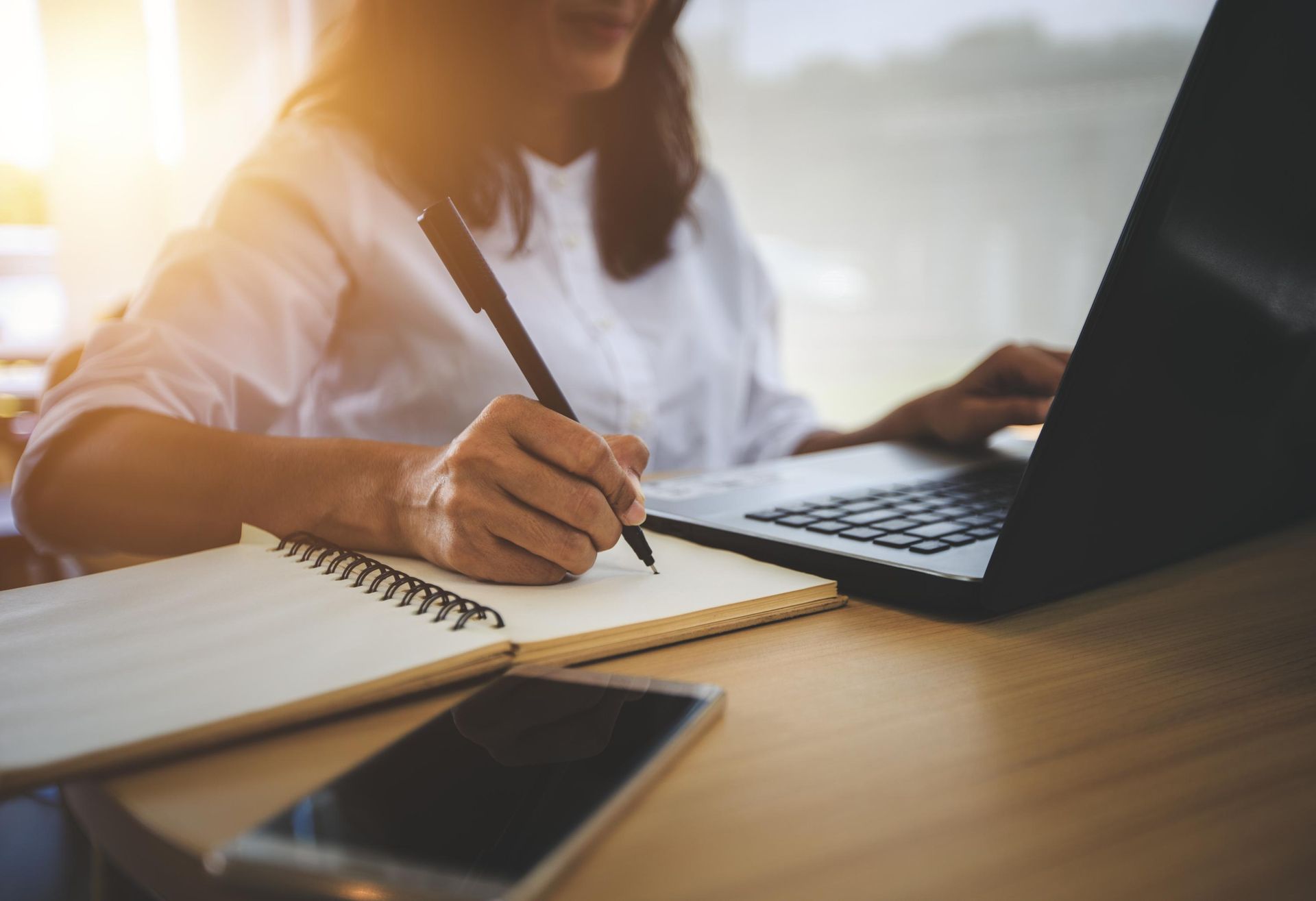 A woman is writing in a notebook while using a laptop computer.