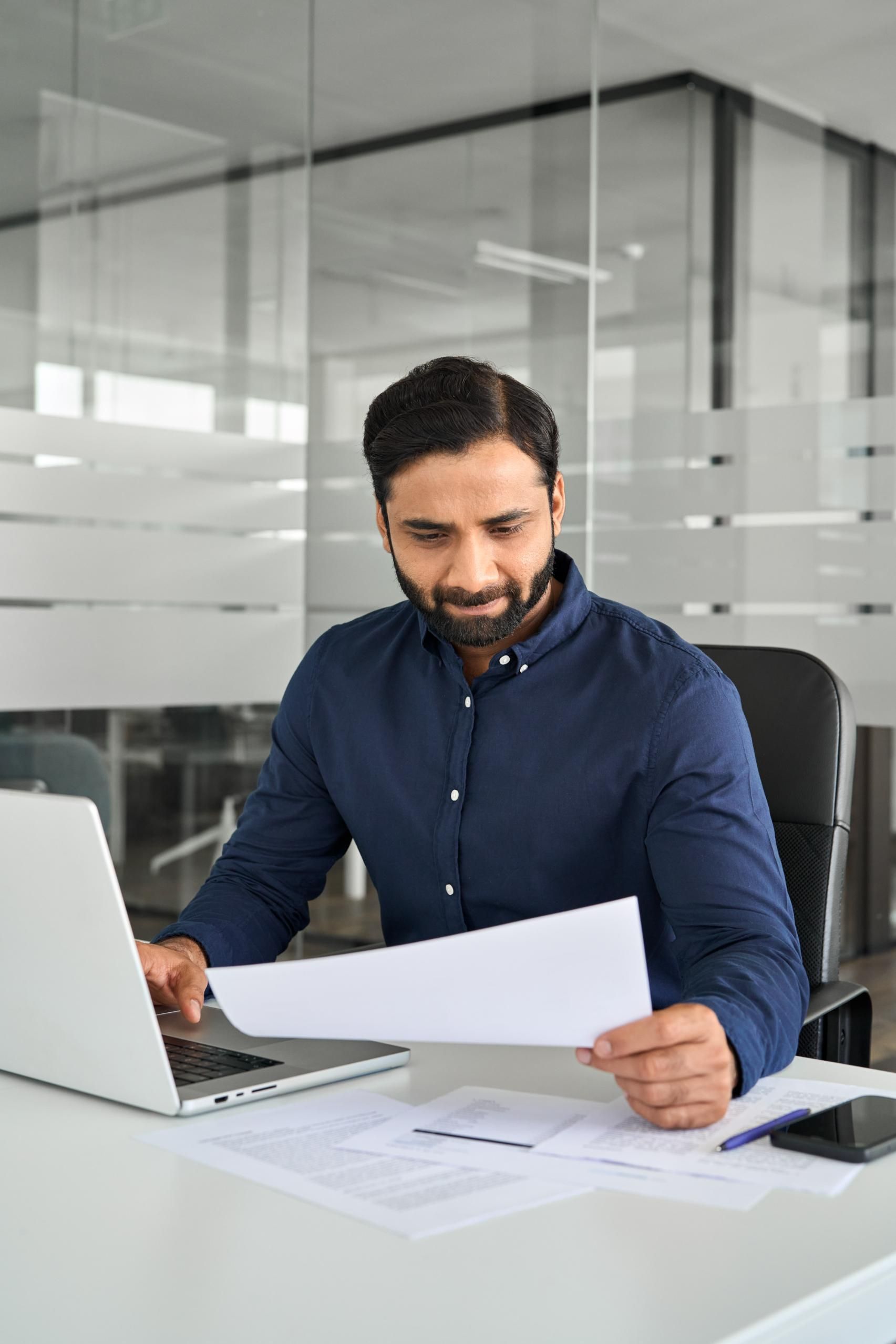 A man is sitting at a desk in front of a laptop computer holding a piece of paper.