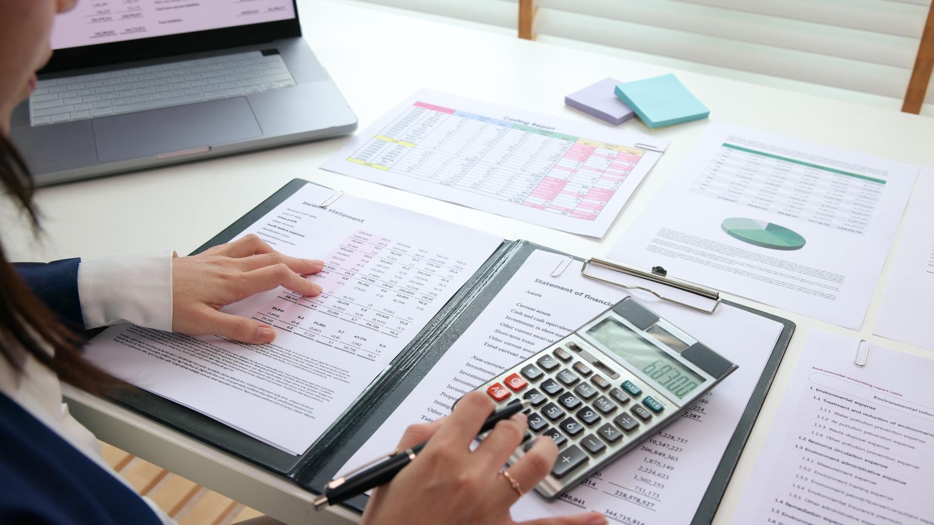 A woman is sitting at a desk using a calculator and a laptop.