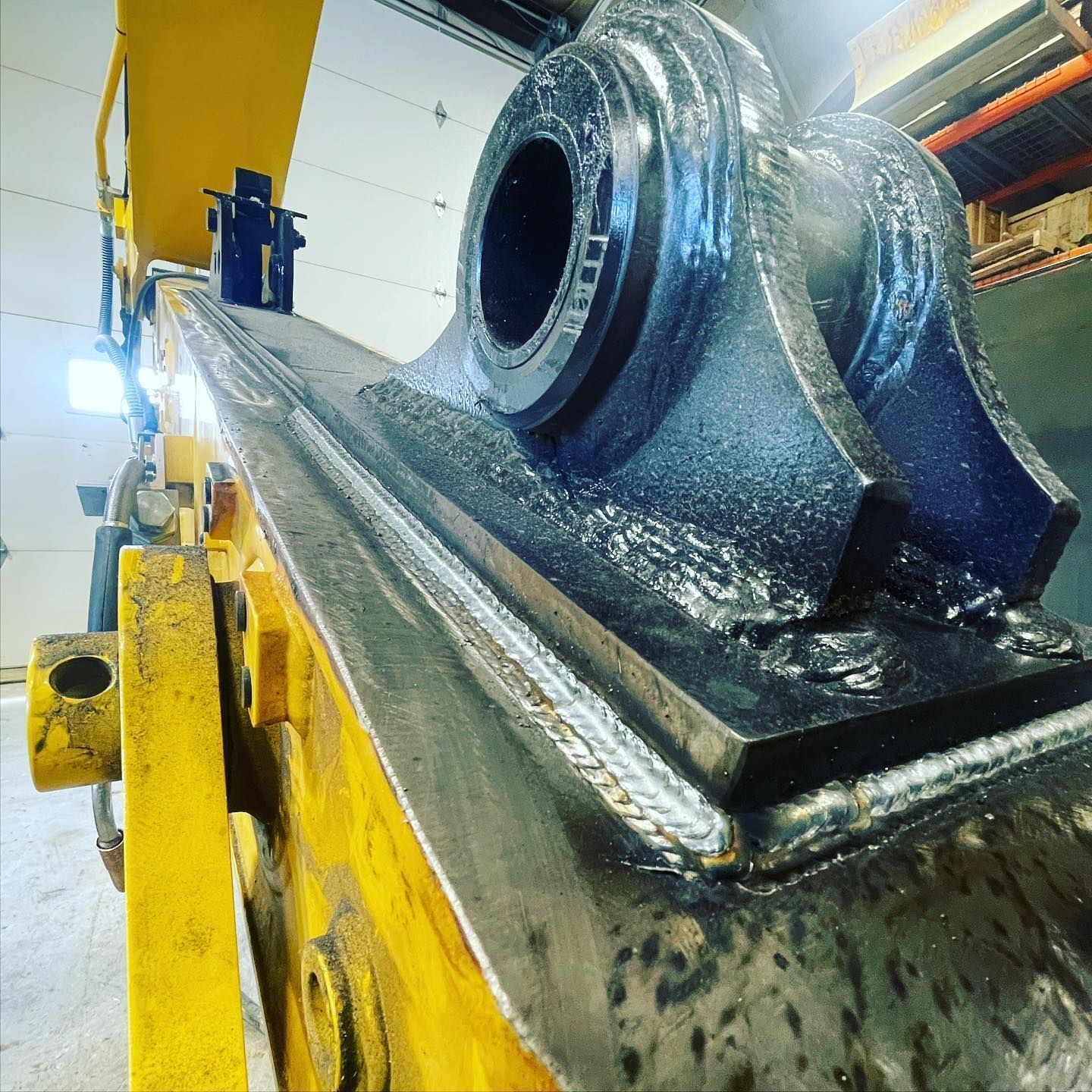 Close-up of a freshly welded black steel pivot bracket attached to a yellow industrial excavator arm in a workshop.