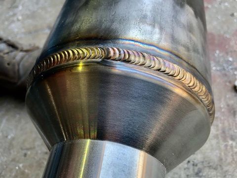 A close-up view of a high-quality circular weld connecting two metal pipes, showing heat discoloration and bead texture.