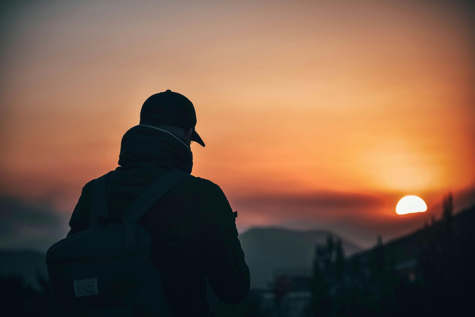 A person with a backpack and cap stands in silhouette against a vibrant, orange sunset over distant hills.