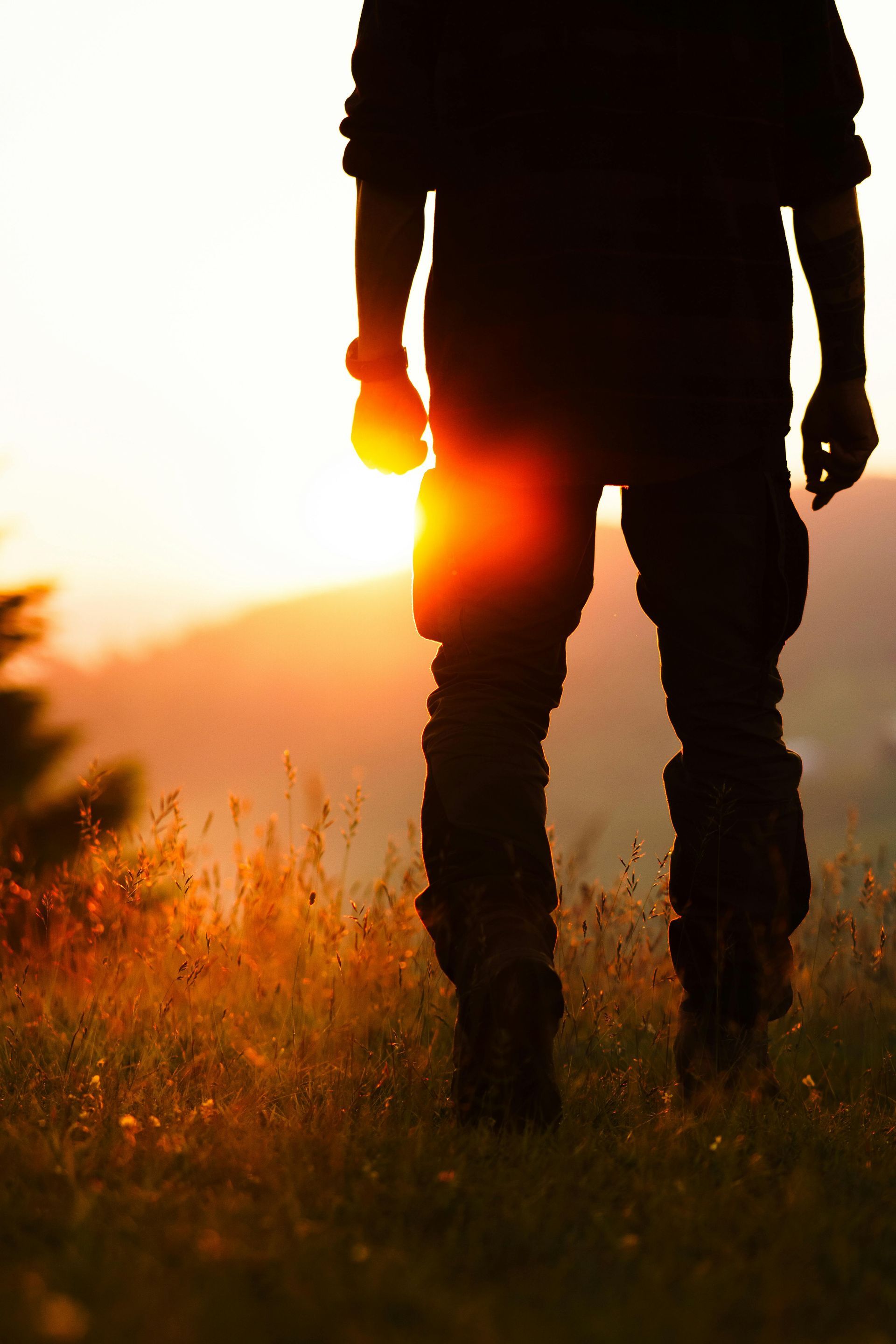 A person in outdoor gear stands in a grassy field at sunset, illuminated by warm, golden backlight.