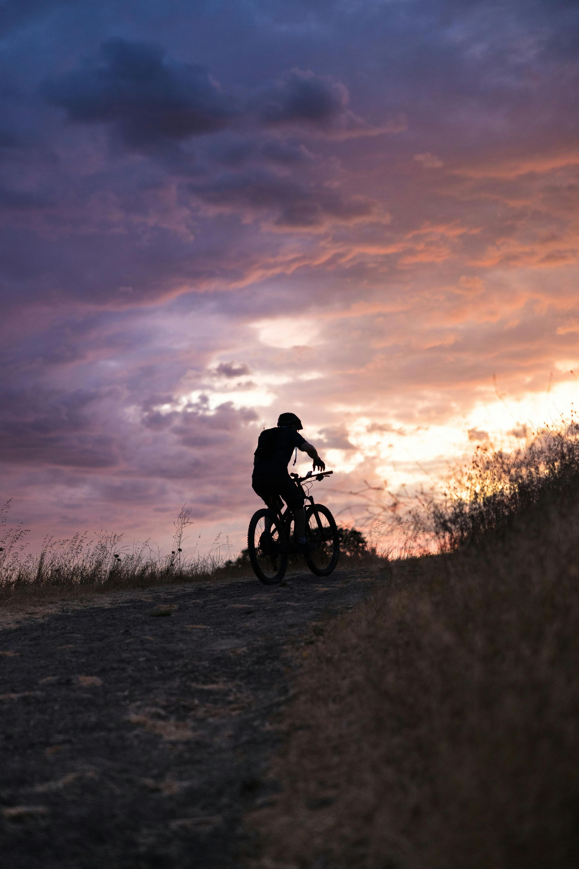 A cyclist rides a bike on a dirt trail at sunset, silhouetted against a cloudy, orange and purple sky.