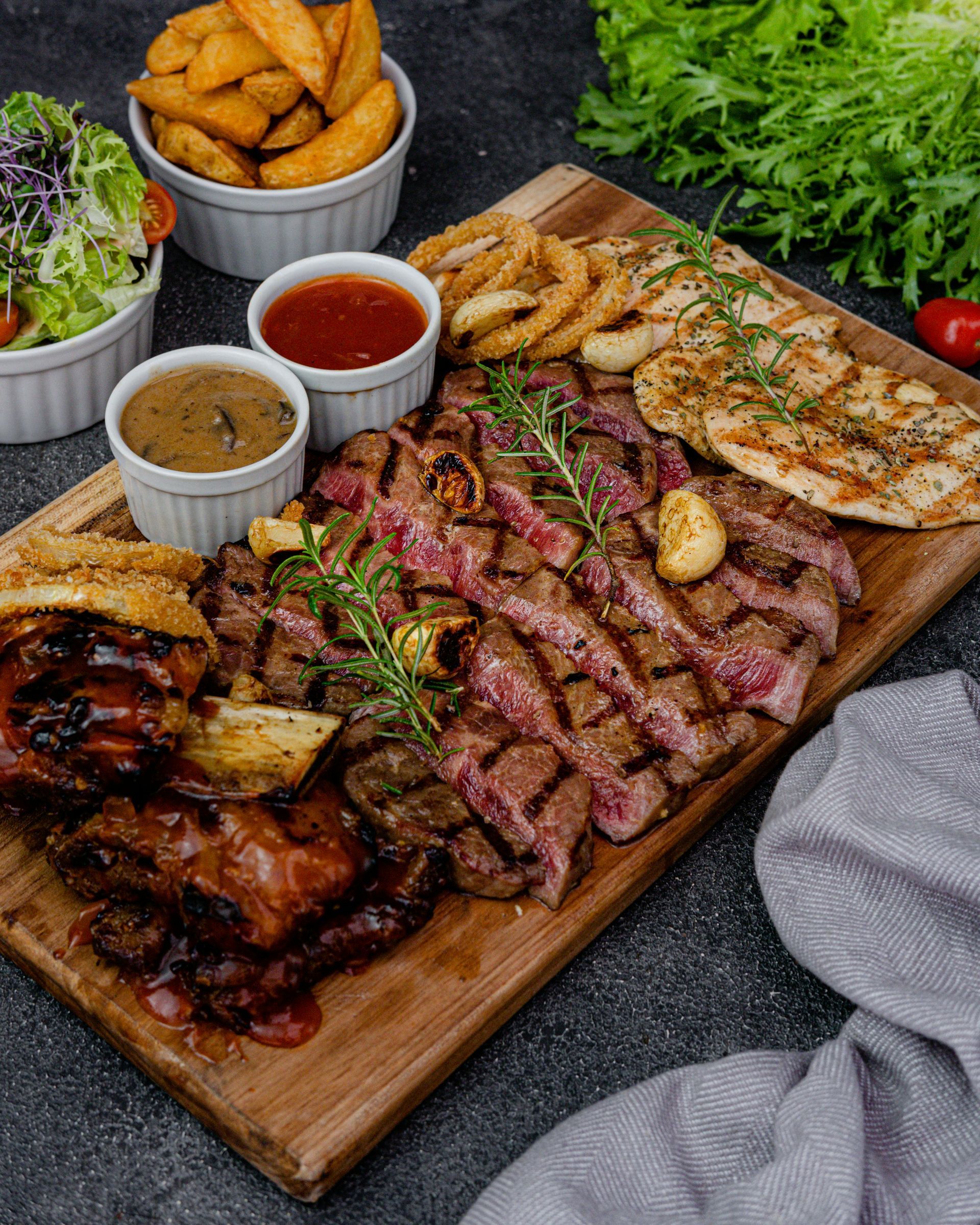 A wooden platter features sliced steak, grilled chicken, onion rings, and sauces, served with side salads and potato wedges.