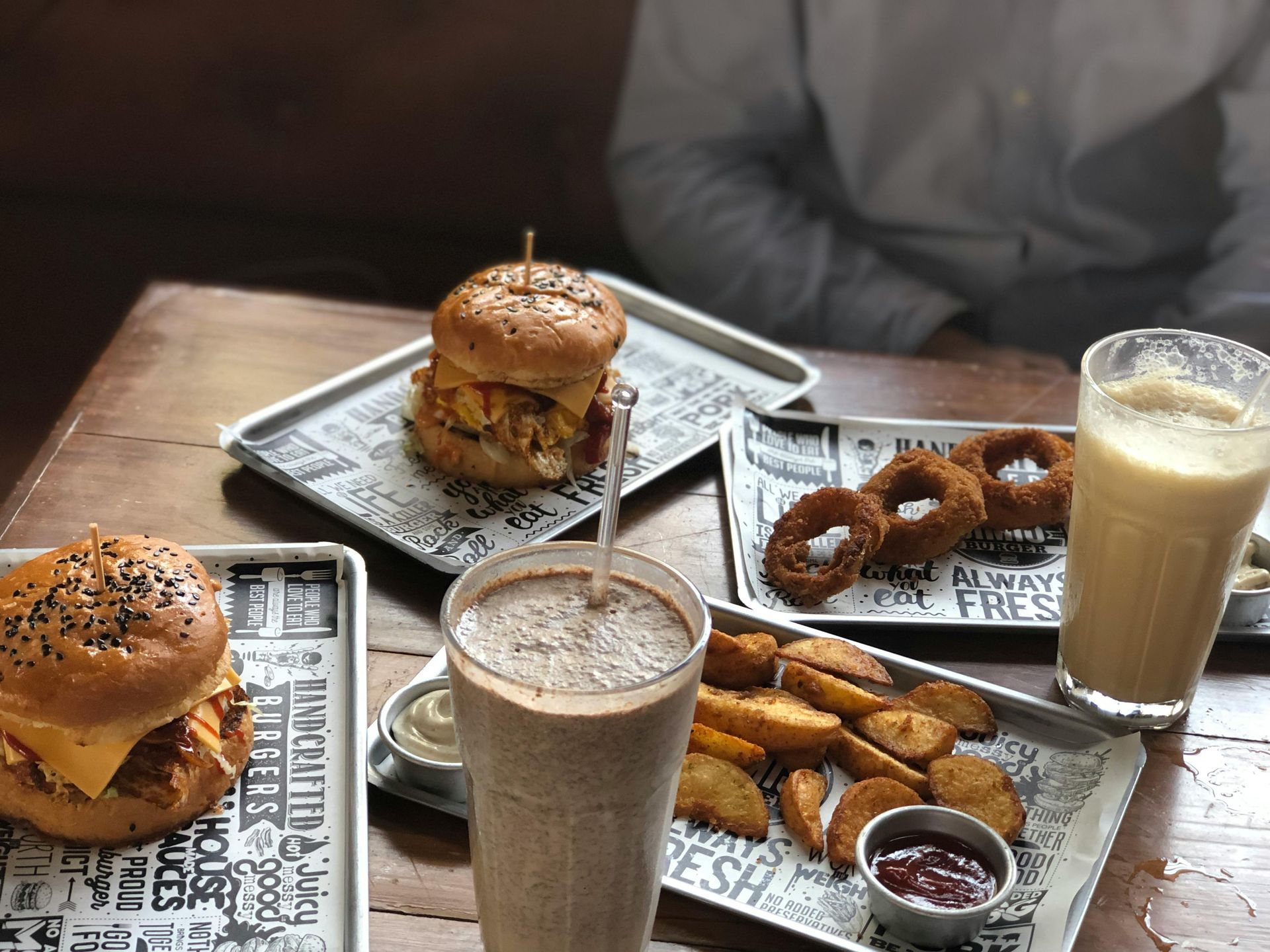 A wooden table set with burgers on trays, onion rings, wedges, and two milkshakes in a restaurant.