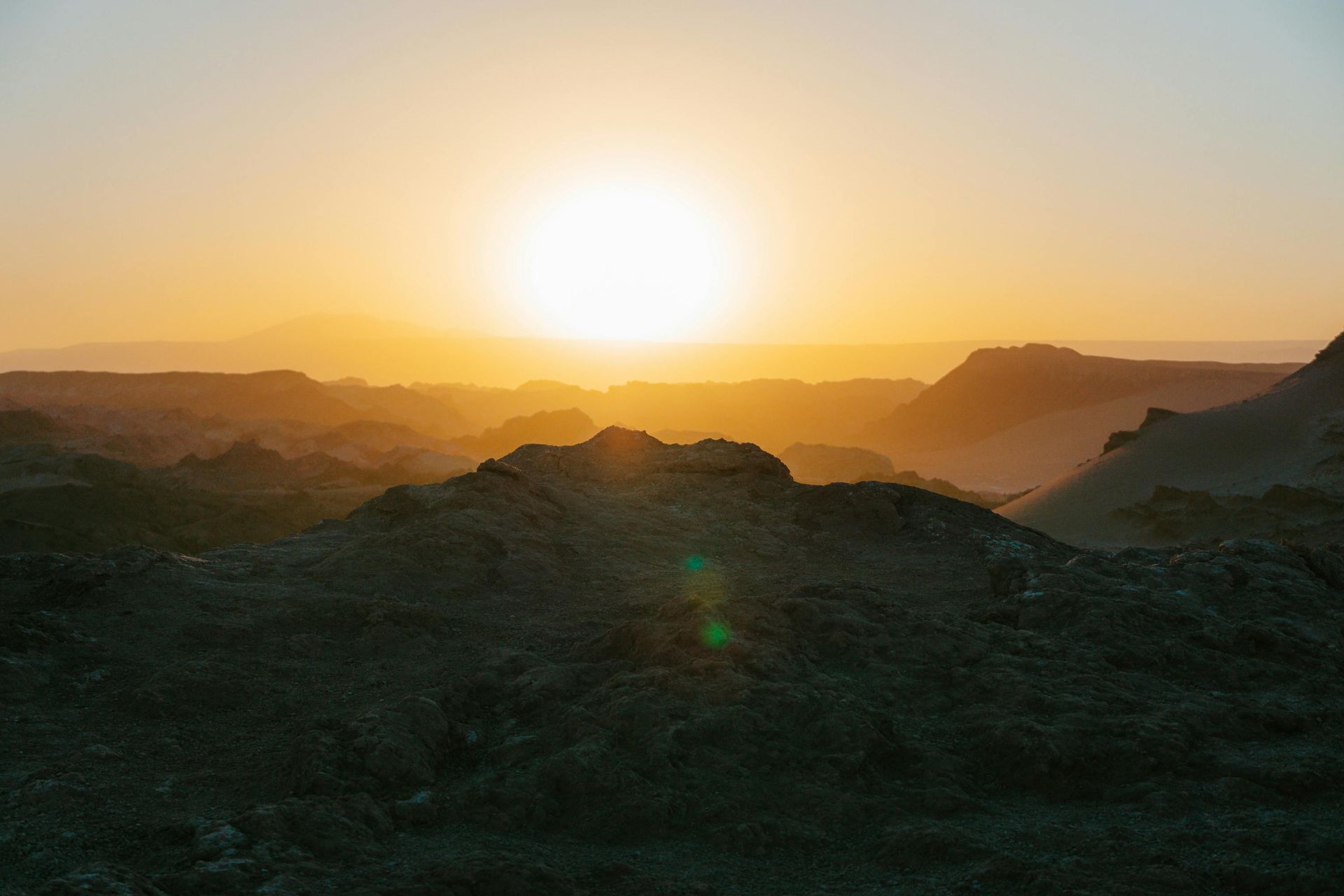 A bright sun sets over a desert landscape with rugged, shadowed hills and a golden, hazy horizon.