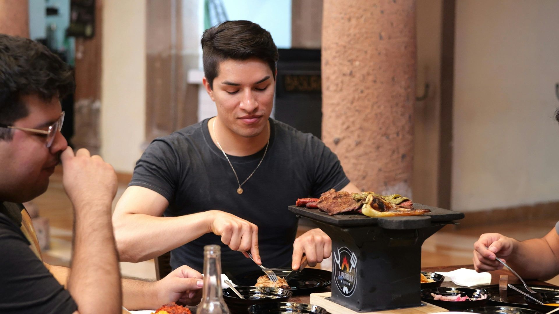 People at a table eat grilled meat from a small, black stone tabletop grill in an indoor setting.