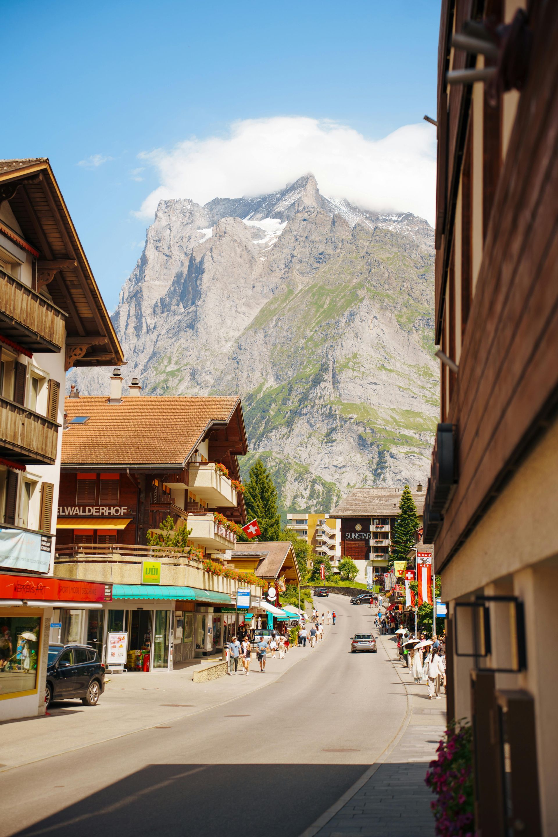 A street in a European alpine village with wooden buildings, shops, and a towering, rocky mountain peak in the distance.