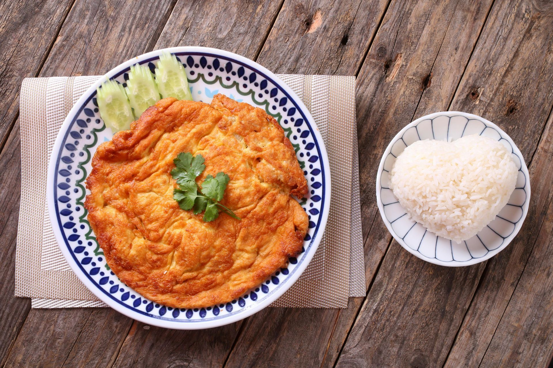 A crispy, golden-brown Thai-style omelet garnished with cilantro, served with sliced cucumber and heart-shaped rice.