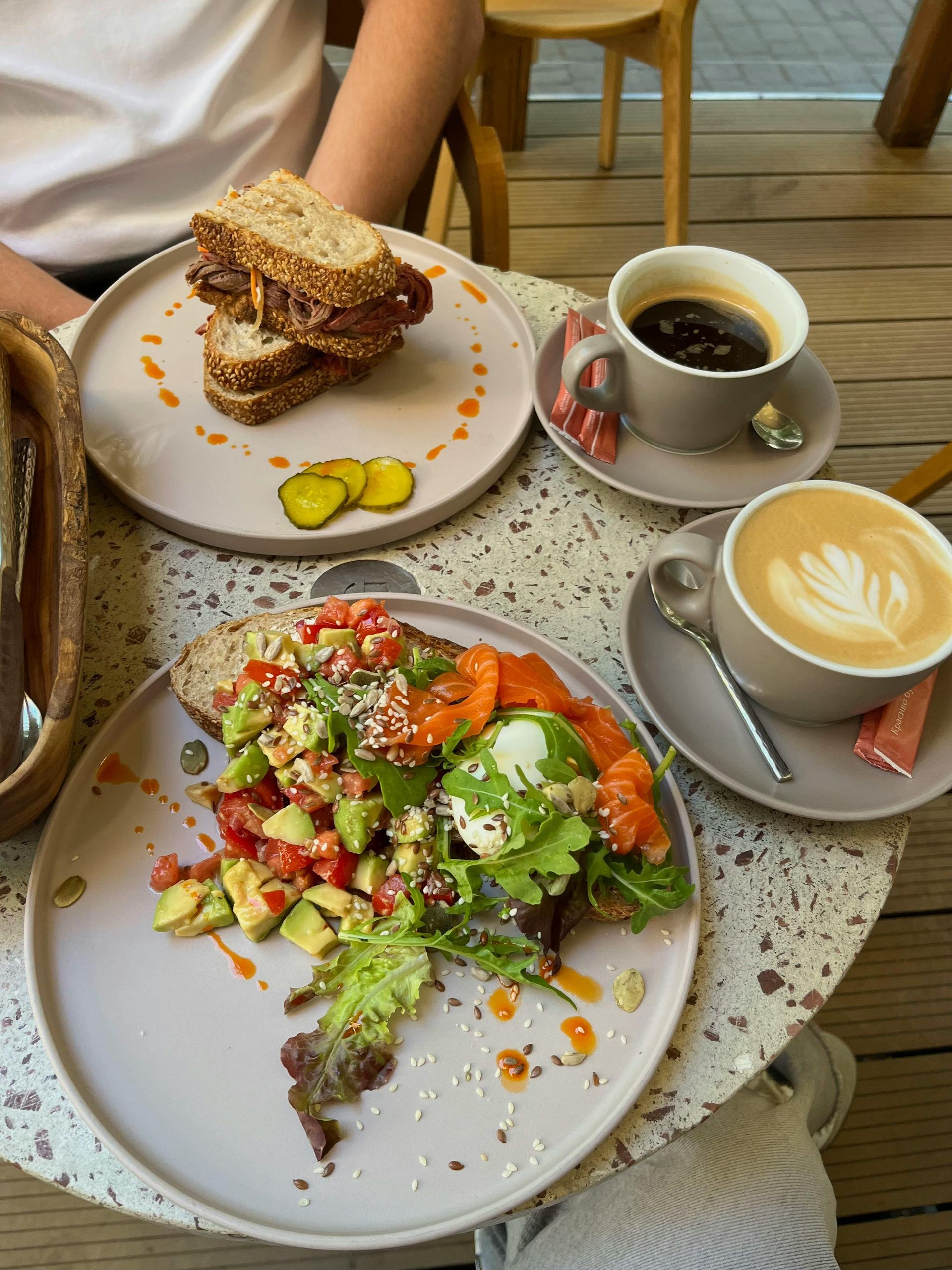 Two plates with a sandwich and avocado toast with salmon, accompanied by a cup of black coffee and a latte on a table.