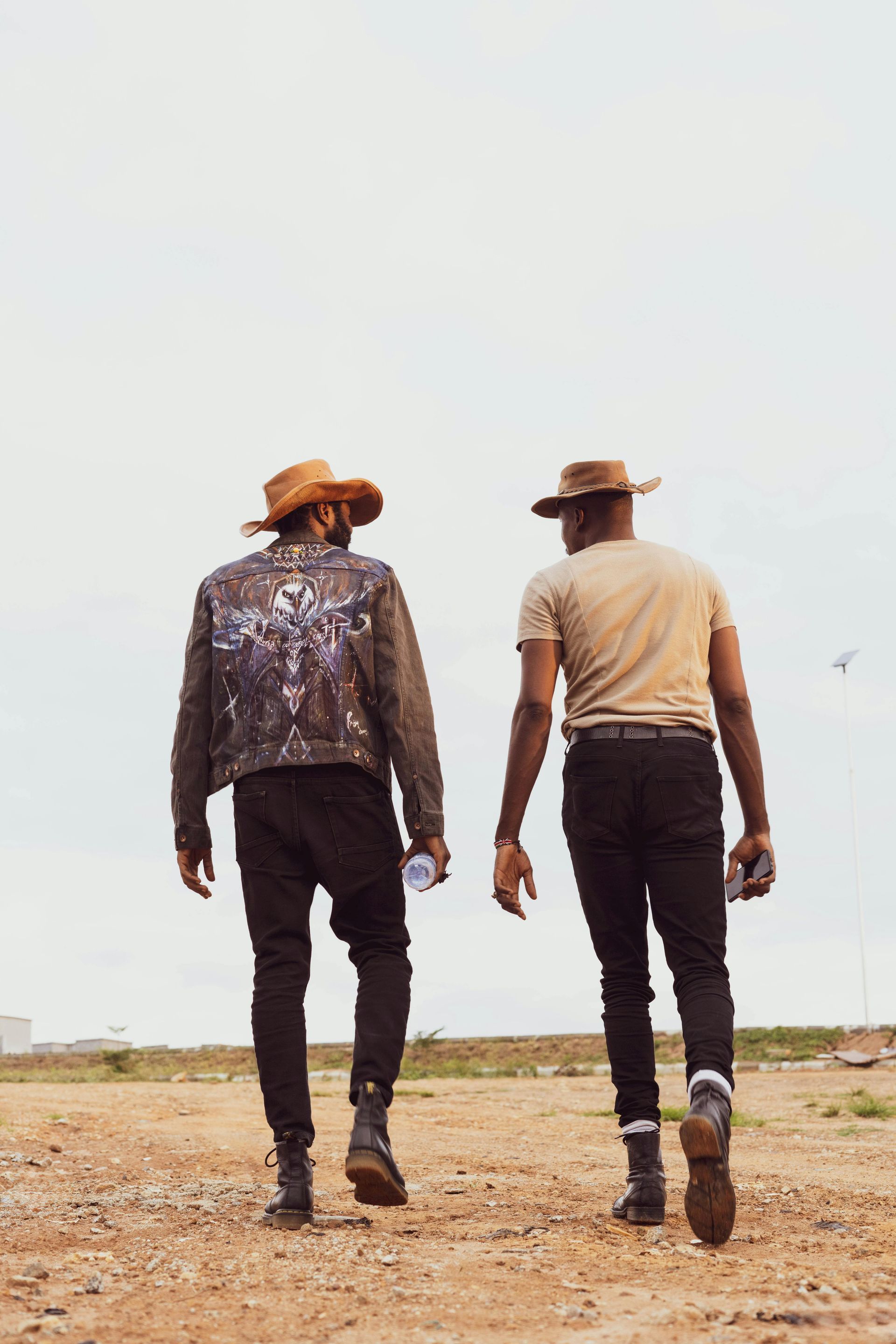 Two people in cowboy hats and dark clothing walk away from the camera across a dry, open landscape under a bright sky.