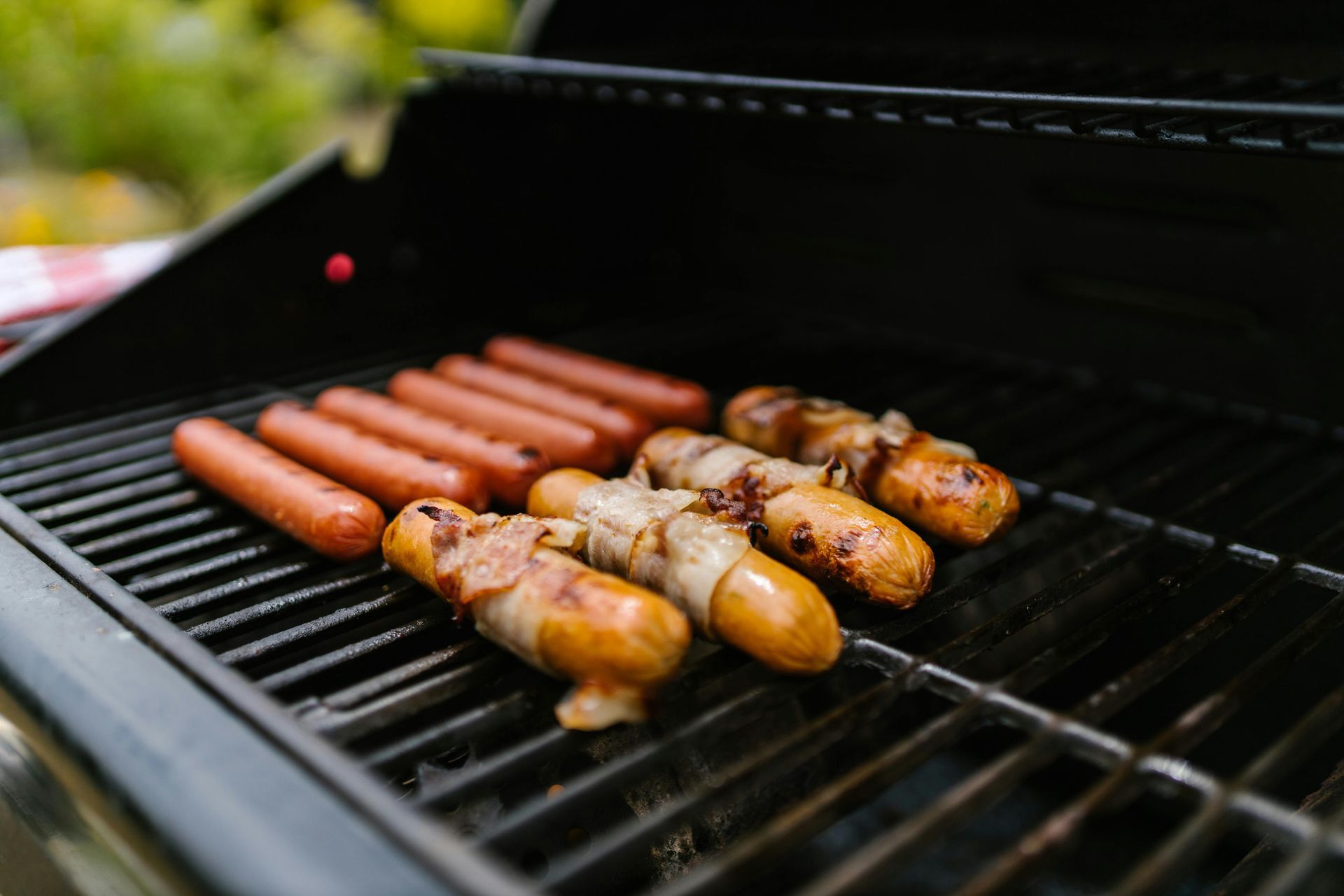 Hot dogs and bacon-wrapped sausages cooking on a black outdoor grill grate.