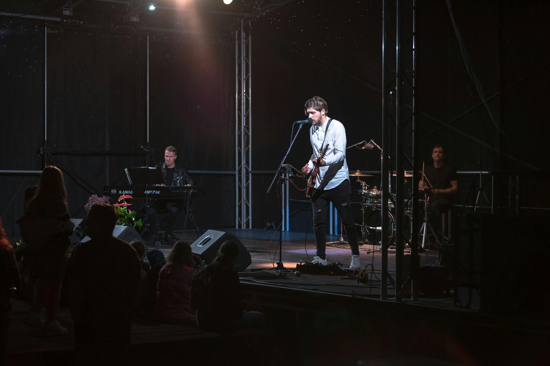 A band performs on a dimly lit outdoor stage, with a guitarist singing into a microphone, a keyboardist, and a drummer.