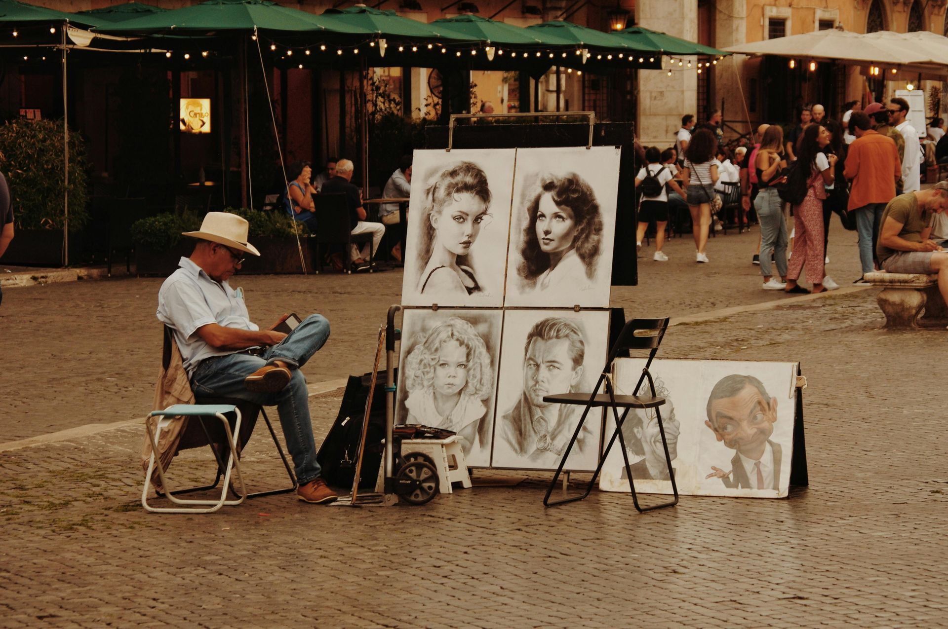 An artist sits on a folding chair in a plaza beside an easel displaying charcoal portraits and a caricature.