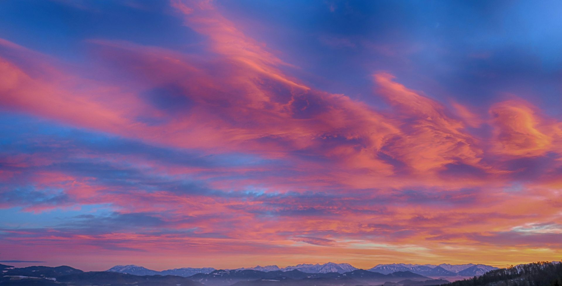 Vibrant pink and orange clouds drift across a deep blue sky at sunset above a misty, dark mountain horizon.