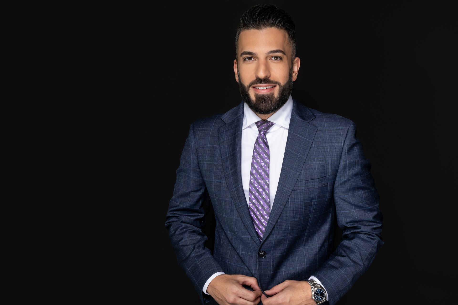 Man in blue suit and patterned tie, smiling against black backdrop.