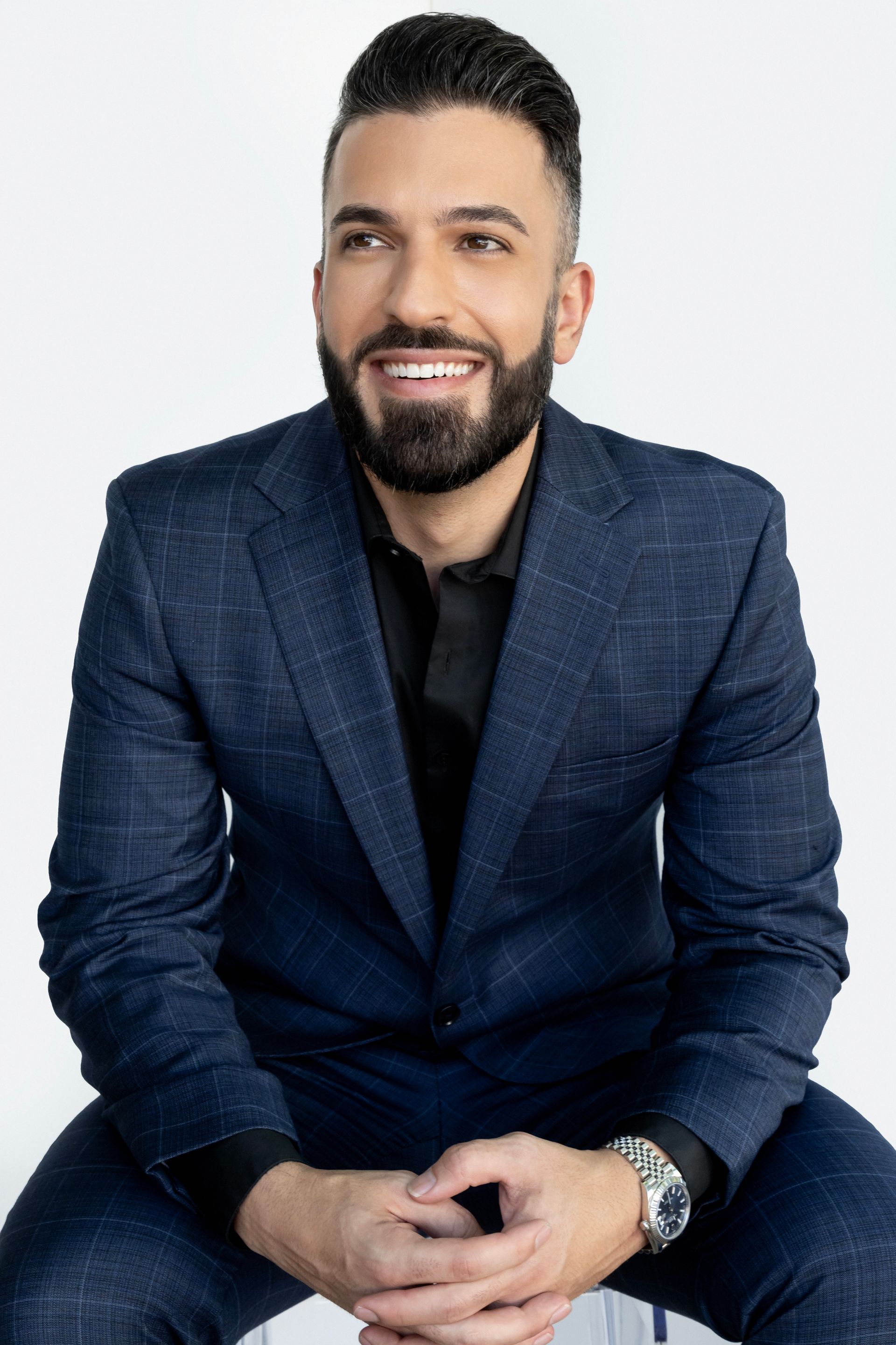 Man in a navy suit, black shirt, and watch, smiling, sitting against a white background.