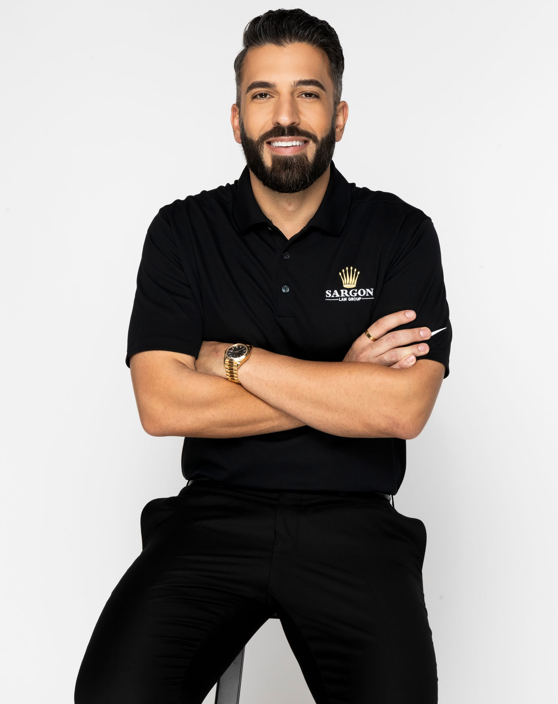 Man with a beard in black polo, crossed arms, sitting on a stool, smiling, studio setting.
