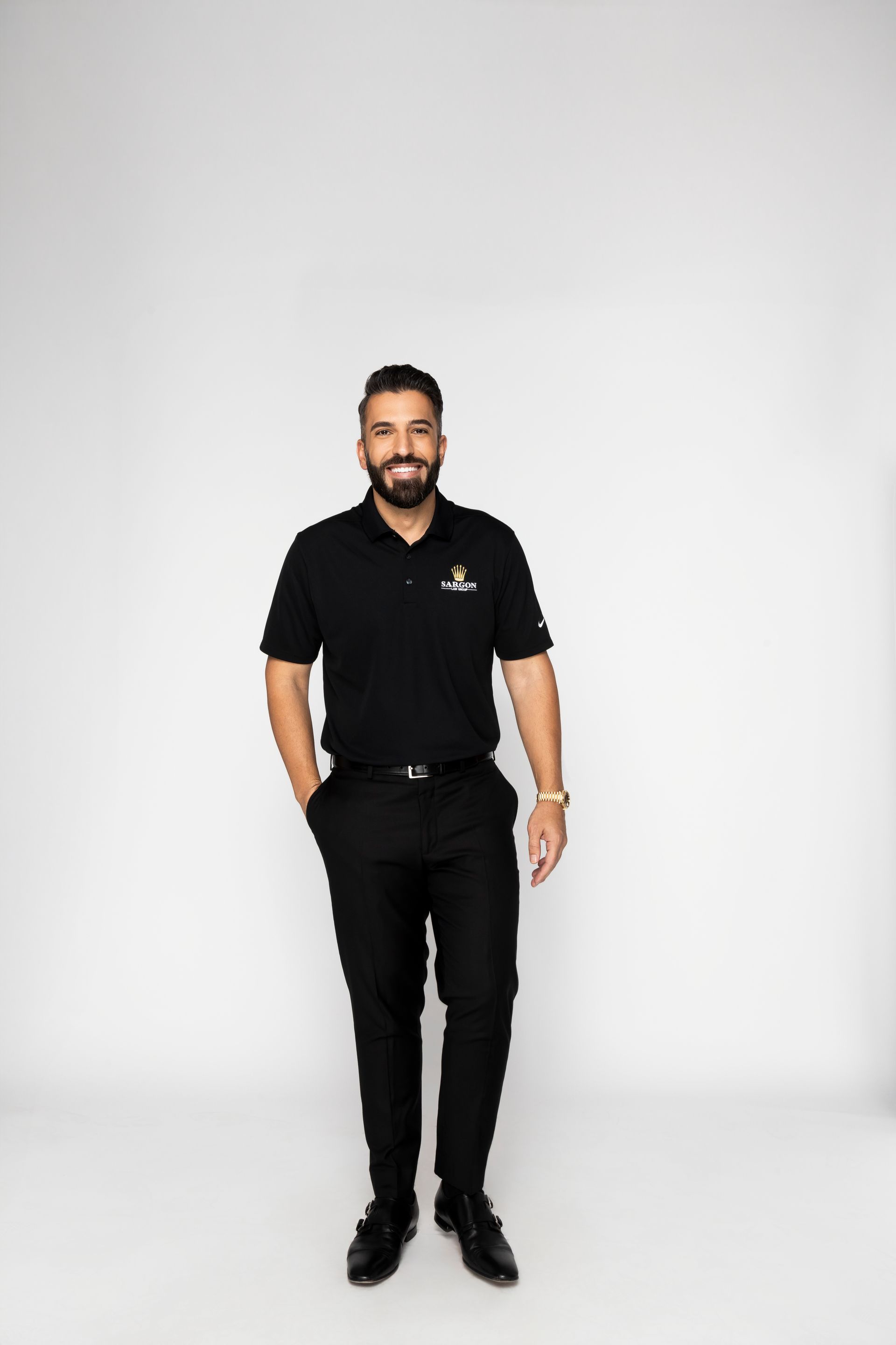 Man in black polo and pants smiling, hands in pockets, standing against white background.