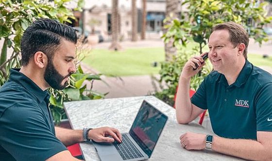 Two men are sitting at a table with a laptop and a cell phone.
