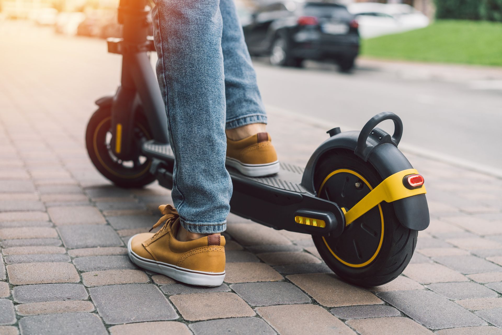 Person riding a black electric scooter on a brick sidewalk; yellow accents, sunny day.
