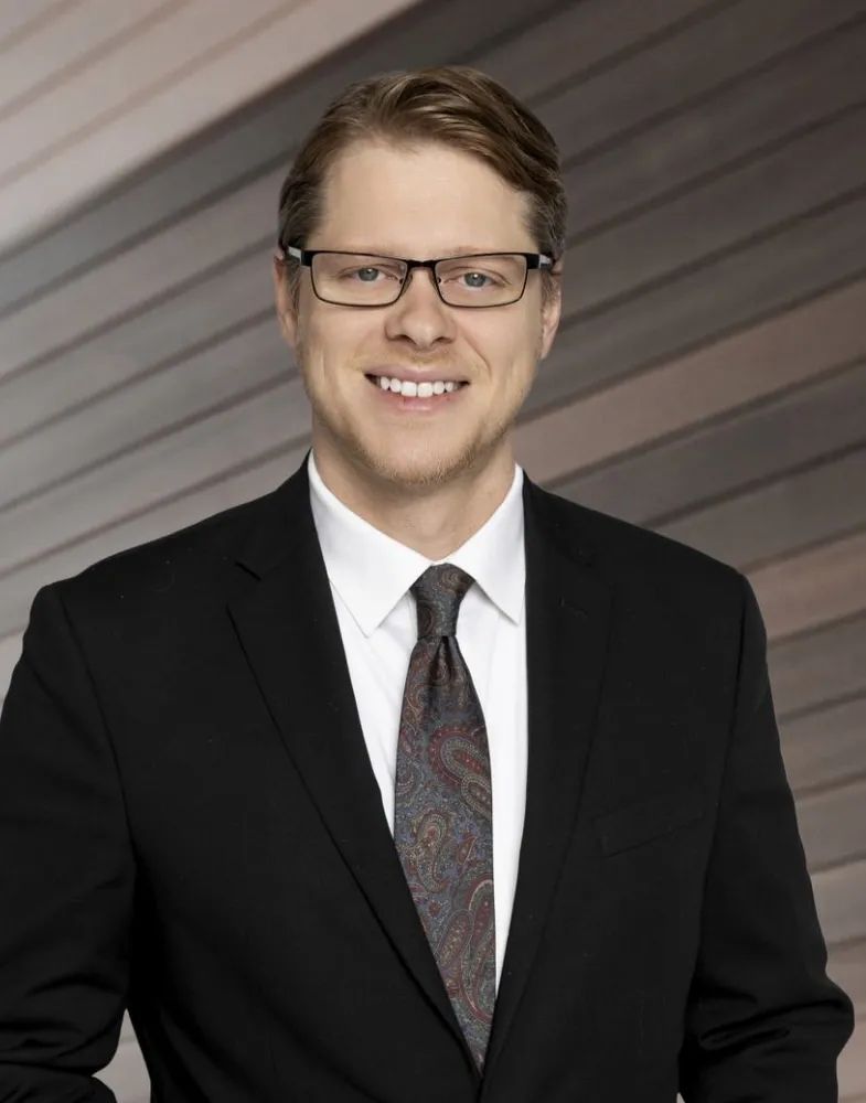 Man in a black suit, tie, and glasses smiles at the camera against a wood-paneled wall.