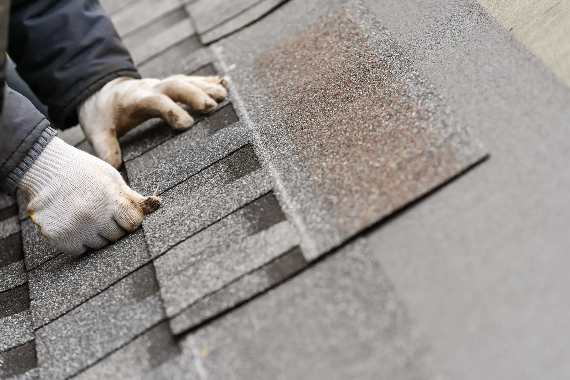 Close-up of a worker in gloves installing asphalt roofing shingles on a residential roof.