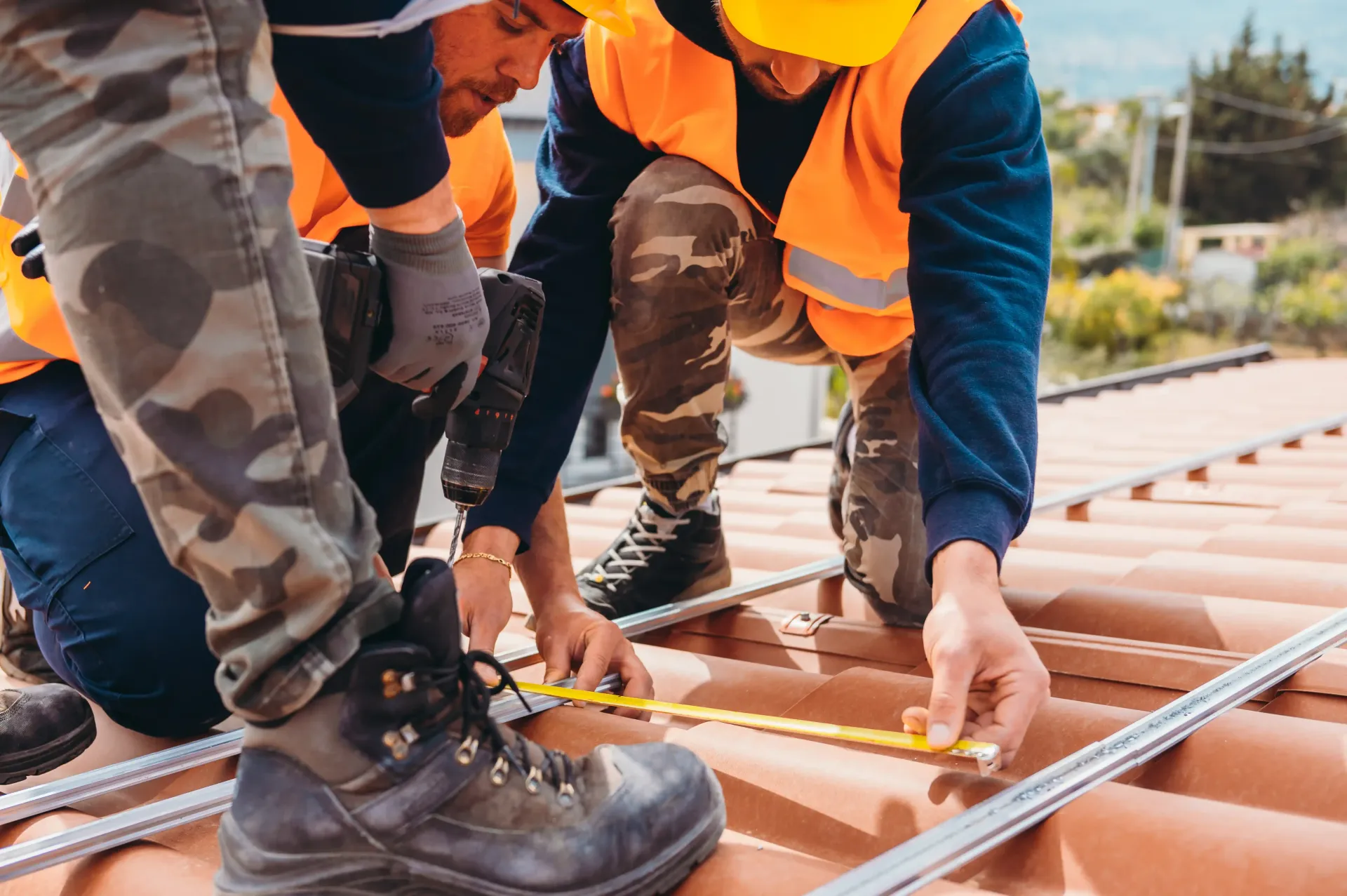 Two construction workers wearing safety vests use a tape measure on a residential tile roof while installing metal rails.
