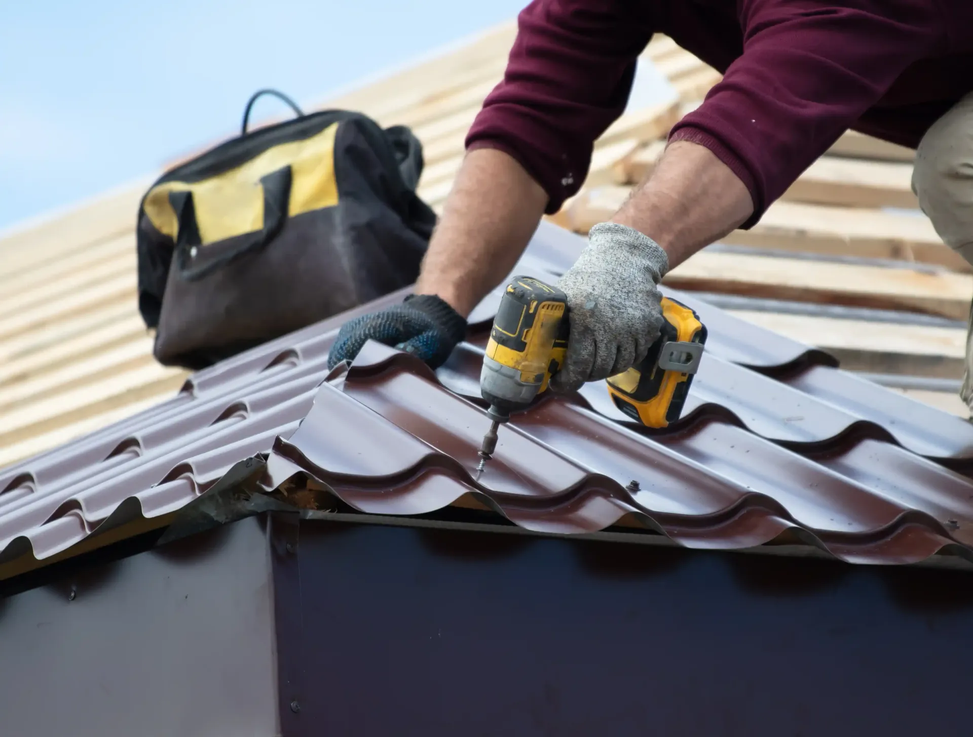 A worker in work gloves uses a drill to secure brown metal roofing panels onto a wooden roof frame with a tool bag nearby.