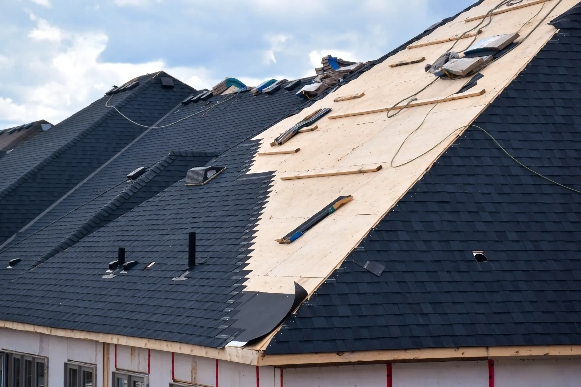 Roofing work in progress on a multi-unit building with dark shingles installed and some plywood sheathing exposed.