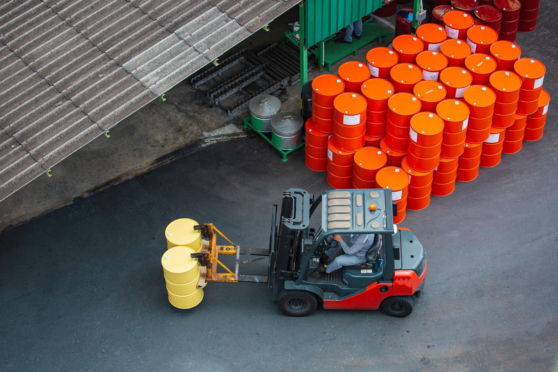 Forklift carrying two yellow barrels near a warehouse, stacks of orange barrels in the background.