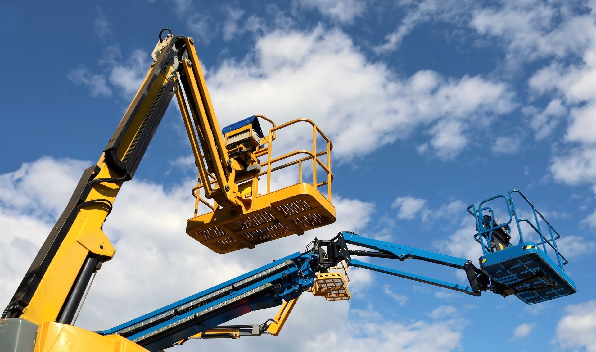 Two elevated boom lifts against a blue sky, one yellow, one blue.