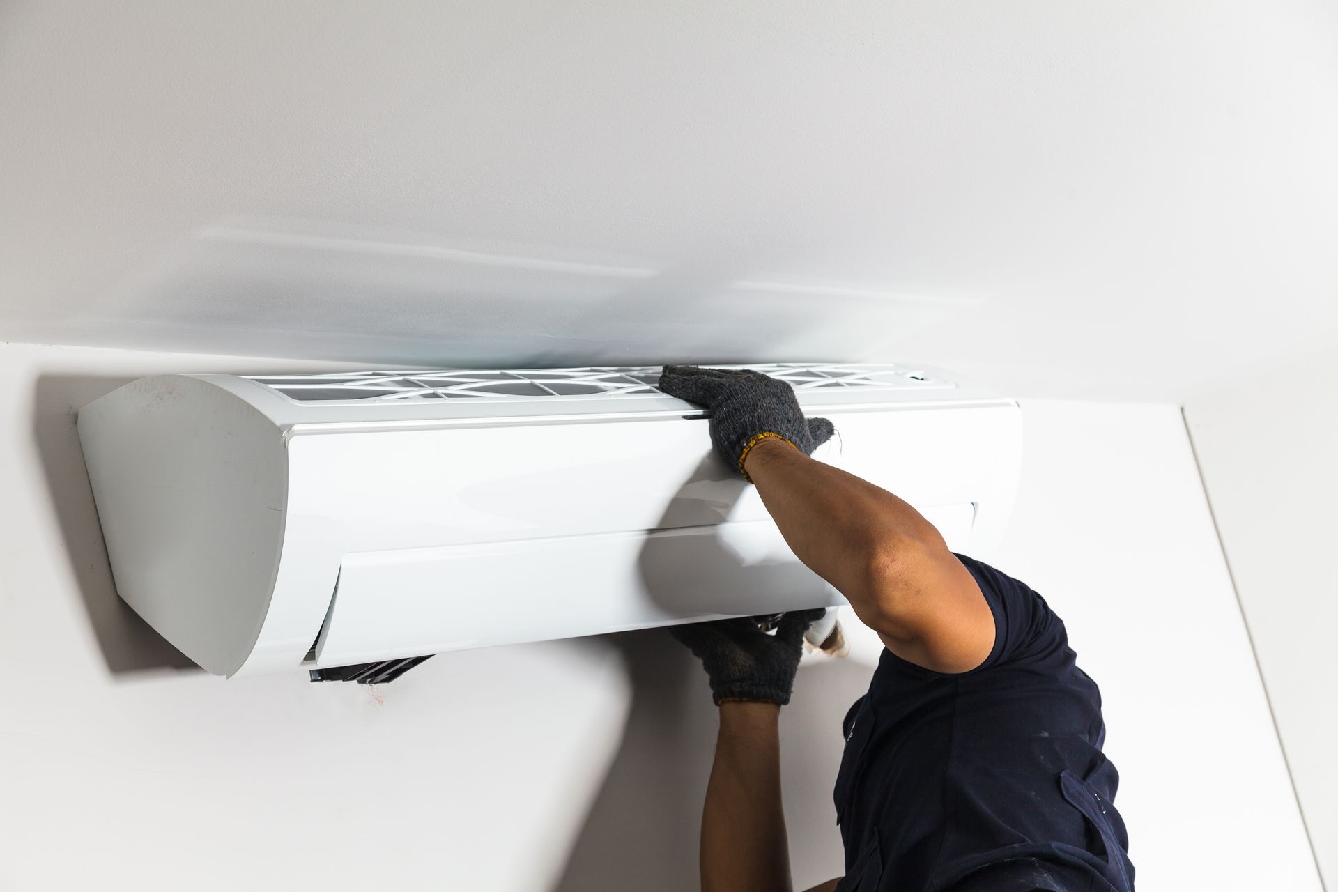 A person in a dark shirt and gloves installs a white air conditioning unit on a plain white wall.