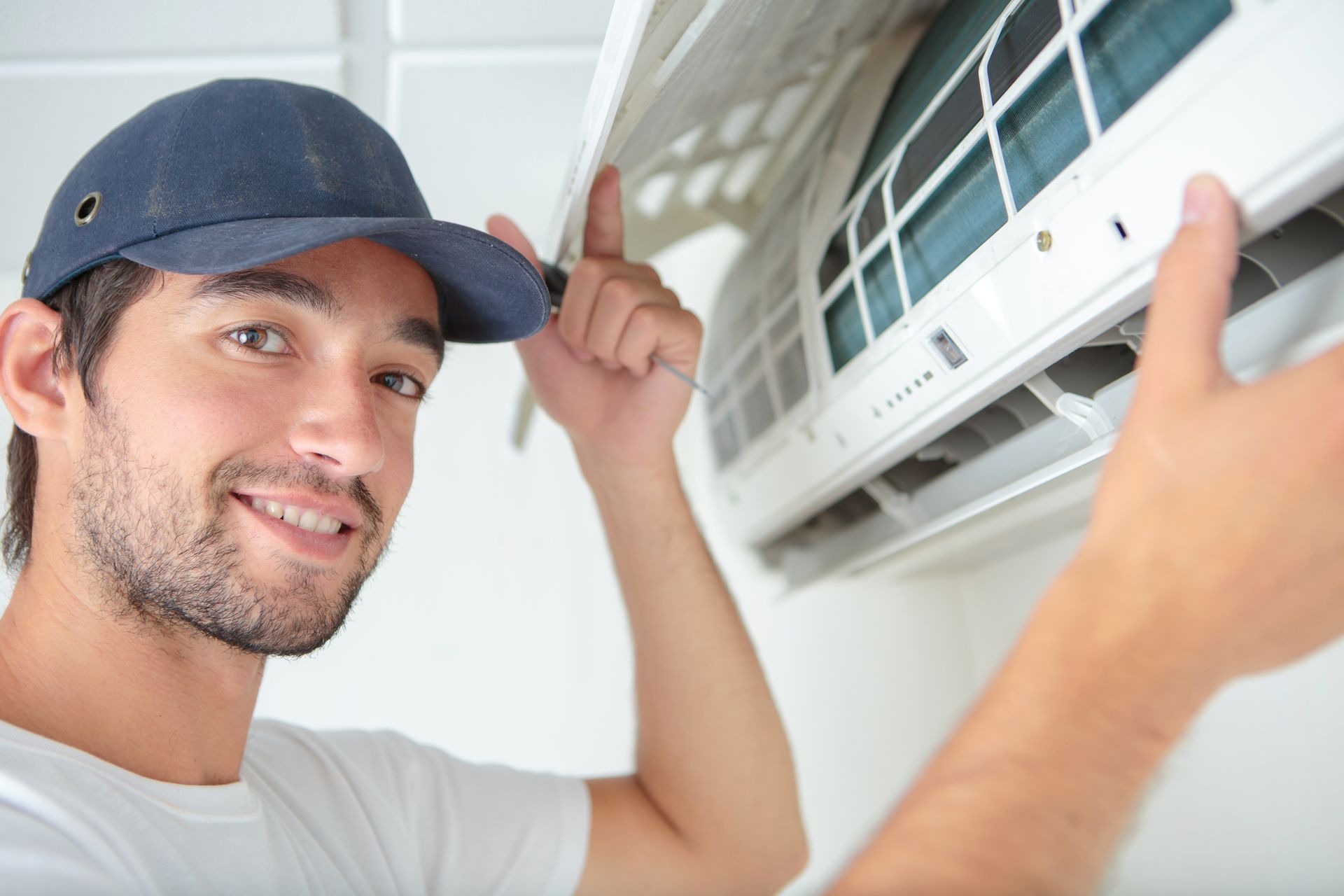 A technician in a blue cap smiling while using a screwdriver to repair a wall-mounted air conditioning unit.