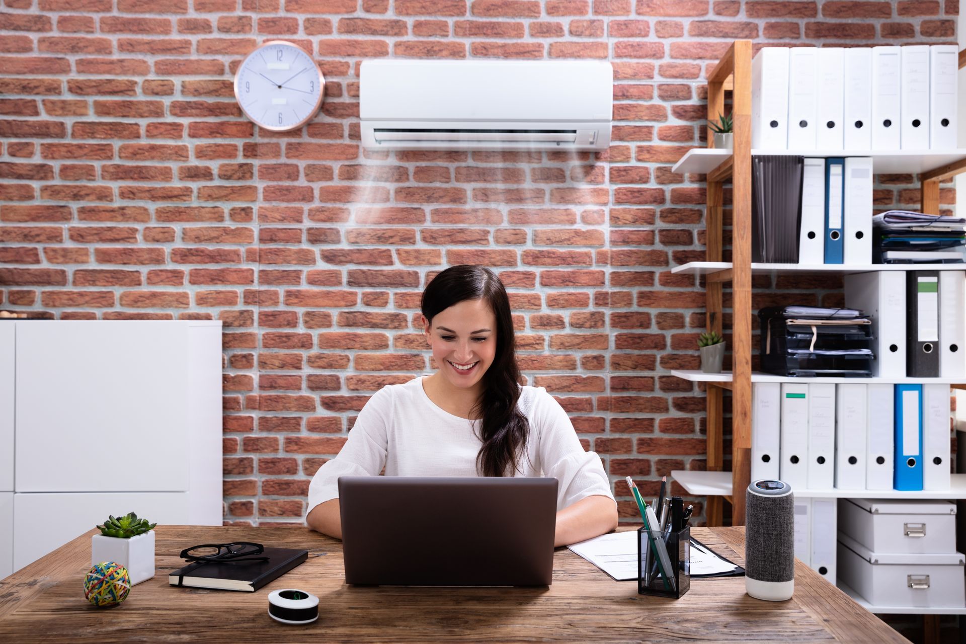 A smiling person works on a laptop at a wooden desk in an office with a brick wall and air conditioner.