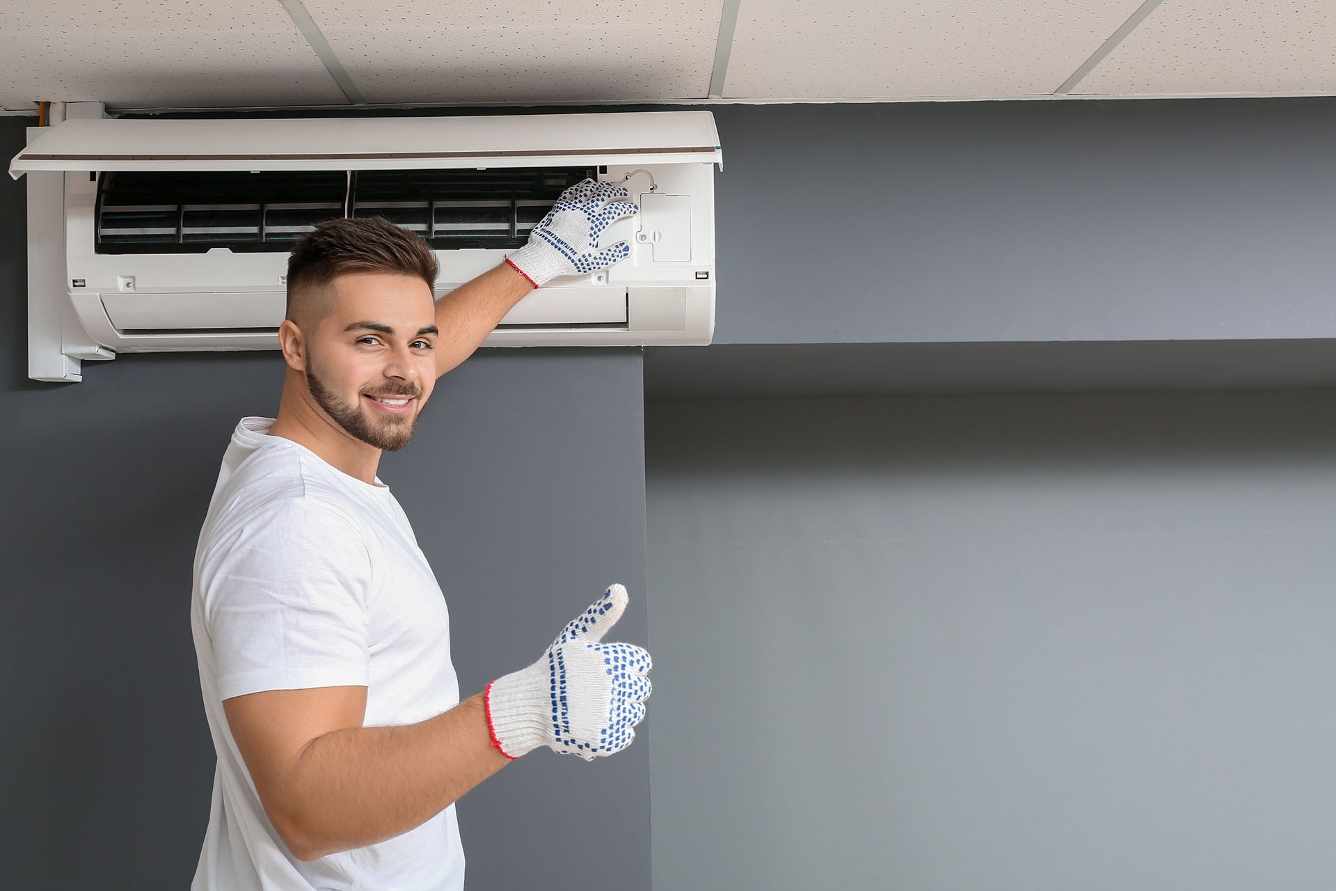 Technician wearing work gloves gives a thumbs-up while servicing a wall-mounted air conditioning unit.